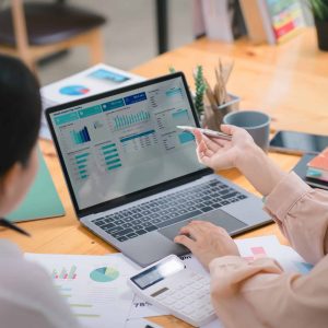 Two businesspeople or an accountant team are analyzing data charts, graphs, and a dashboard on a laptop screen in order to prepare a statistical report and discuss financial data in an office.